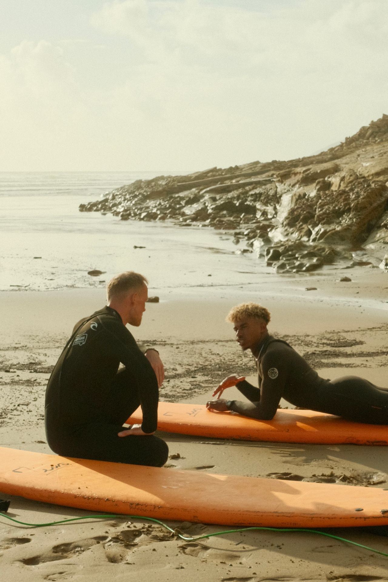 Two surfers in wetsuits on orange surfboards, talking on a sandy beach with rocky shoreline.